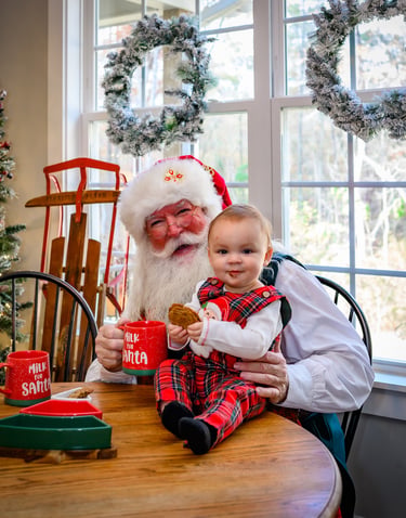 Santa Claus sits at a wooden table with a baby in plaid pajamas, enjoying cookies and mugs of milk.