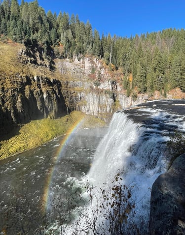 Mesa Falls cascading into a canyon with a vibrant rainbow over the water and evergreen forest.