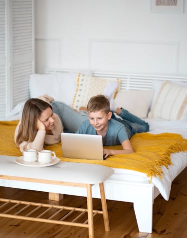 Smiling mother and son lying on a clean white bed, watching something together on a laptop