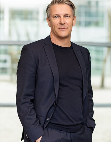 Een man in een donkerblauwe blazer, zwart shirt en donkerblauwe broek. Hij lacht naar de camera