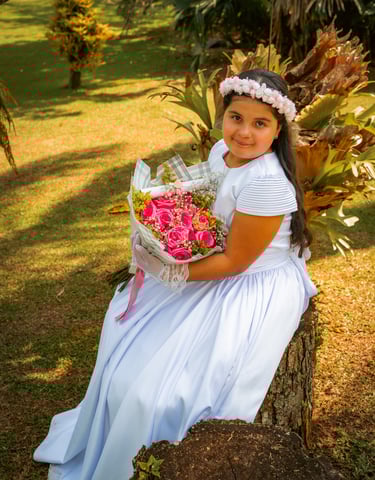 Niña con un vestido de primera comunión blanco sosteniendo un ramo de rosas rosas al aire libre.