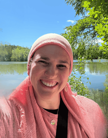Naomi Korbman smiling wearing a pink headscarf in a selfie by a calm lake with lush green trees.