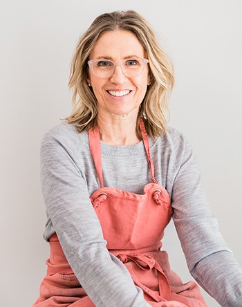 Casual seated portrait with a light gray shirt and pink apron and pink glasses on a plain light background