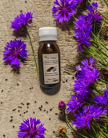 A bottle of Huile de Nigelle black seed oil surrounded by purple cornflowers and organic seeds.