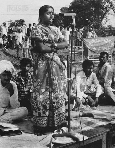 A black-and-white photograph of a woman in a floral sari speaking confidently into a microphone at a public gathering.