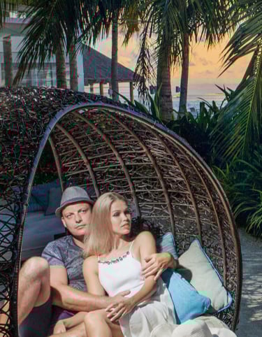 A romantic couple sitting in a cozy, woven wicker pod under tropical palm trees, enjoying
