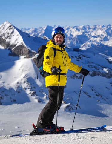 alt= "Xaver Kröll in einer gelben tobe Jacke und schwarzen Hose steht auf einem verschneiten Berg"