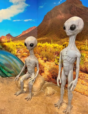 Two grey alien statues at a Roswell UFO exhibit with a desert mountain landscape background.