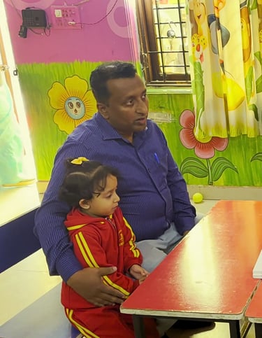 Father and young daughter meeting with a preschool teacher at a small table in a colorful classroom.