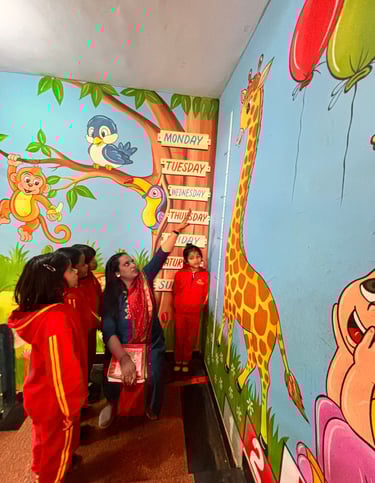 A preschool teacher pointing to a colorful giraffe height chart and days of the week mural in a classroom.