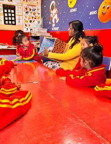 A teacher reads a storybook to preschool students at a red table in a colorful space-themed classroom.