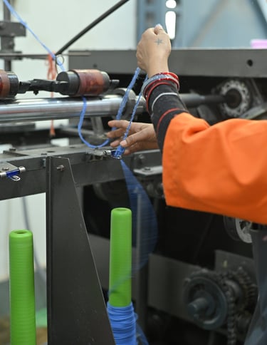 A factory worker feeds blue thread into an industrial textile machine with green spools.
