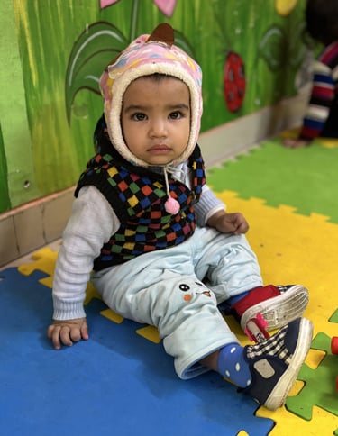 A toddler wearing a warm winter hat sits on colorful foam play mats in a vibrant daycare setting.
