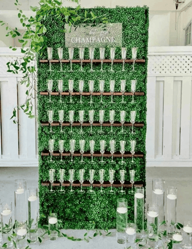 A wall adorned with lush greenery with a "But first Champagne" sign for a wedding.
