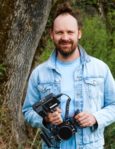 Lächelnder Mann mit Bart und Zopf in blauer Jeansjacke, draußen, Kamera Rig in den Händen, Porträt