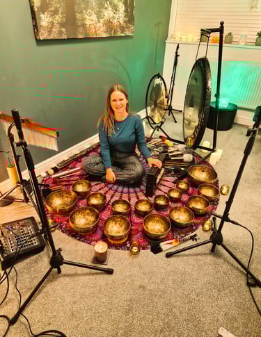 Chrissy sitting on a mat with shruti box, chimes, gongs, many singing bowls, and audio equipment