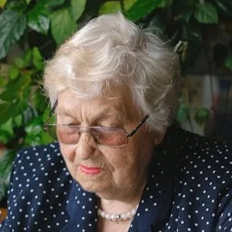 Elderly woman with short white hair wearing glasses and a navy blue polka dot jacket, looking down thoughtfully with greenery