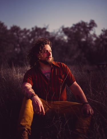 Artistic portrait of a man with long wind-swept hair sits in long grass at sunset
