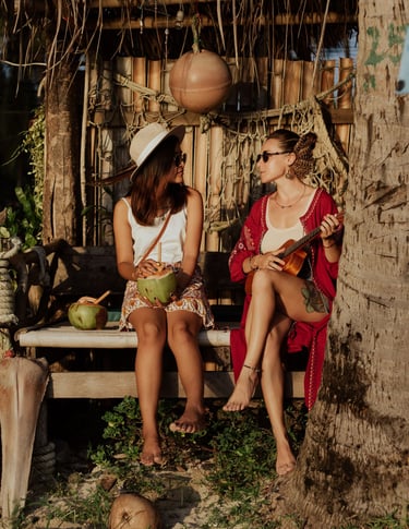 Two women relaxing at a tropical beach bar, drinking fresh coconut and playing ukulele at sunset.
