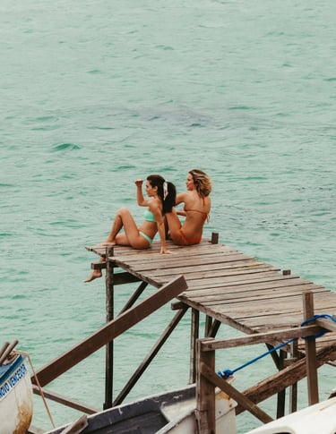 Two women in bikinis sitting on a wooden pier overlooking turquoise tropical ocean water.