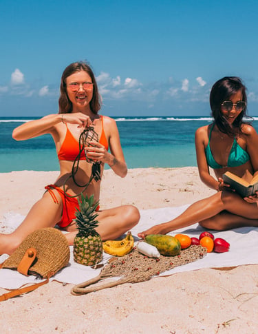 Two young women in bikinis enjoy a tropical beach picnic with fresh fruit and books.