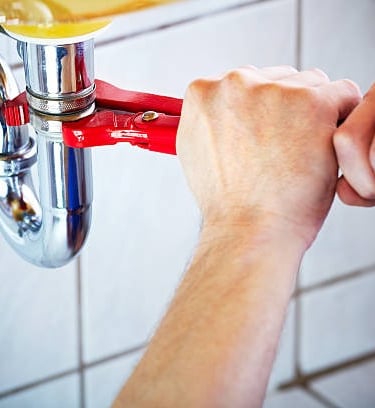 man using a red mini pipe wrench to turn a pipe under the sink