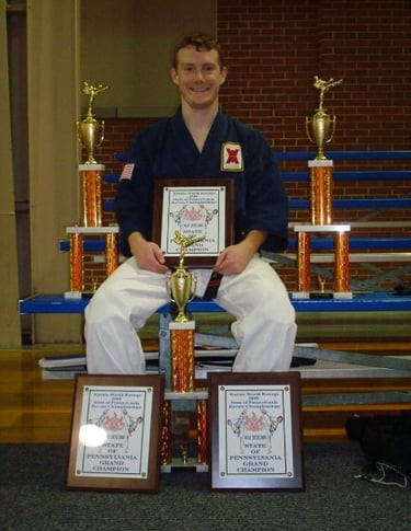 A karate grand champion in a blue gi sits with his martial arts trophies and awards.