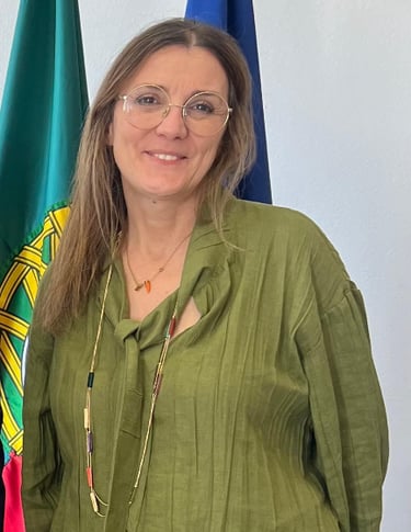A smiling woman with glasses and a green blouse standing in front of the flag of Portugal.
