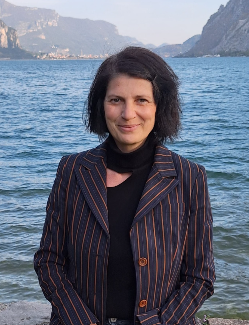 A smiling woman in a pinstripe blazer stands before a scenic mountain lake landscape.