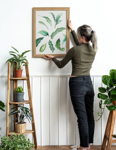 A woman hanging a wooden framed botanical leaf art print on a white wall in a room filled with indoor plants.