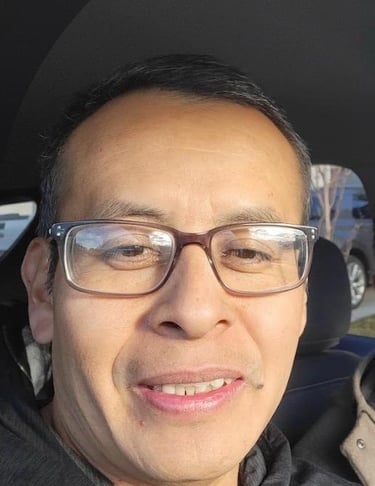 A smiling man wearing rectangular eyeglasses and a black hoodie sitting in a car.