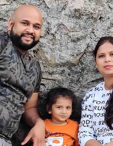 A happy family with a father, mother, and young child posing together in front of a stone wall.