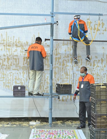 Textile workers on scaffolding hand-weaving a large, intricate gold and white wall hanging.