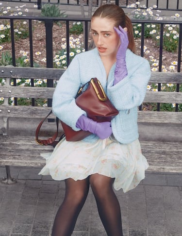 Fashionable woman in a blue tweed jacket and purple gloves holds a luxury leather bag on a park bench.