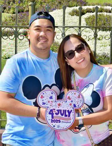 Couple smiling at Disneyland wearing Mickey Mouse shirts and holding a 4th of July 2025 sign.