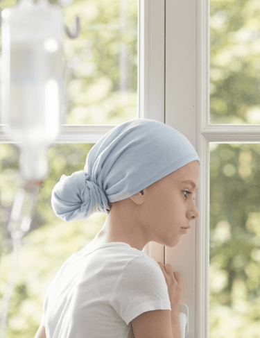 Criança que está passando pelo tratamento contra o câncer, com turbante, olhando pela janela