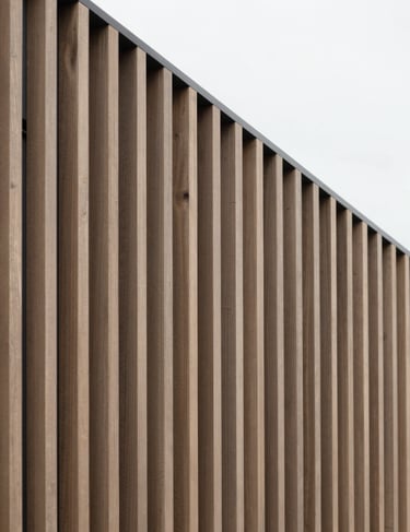 Architectural detail of a facade featuring rhythmic vertical slats in a warm muted wood tone #6B6760 against a crisp #FBF9F6 sky.