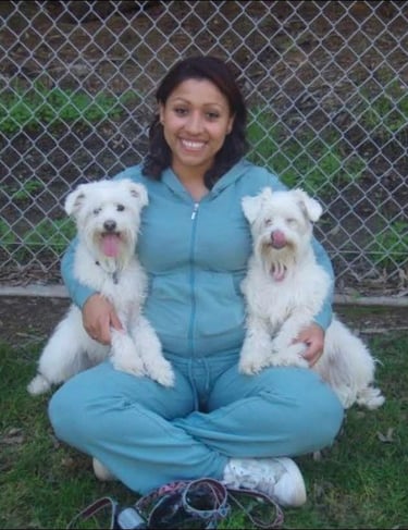 A smiling woman sits cross-legged outdoors holding two fluffy white dogs in front of a chain-link fence.