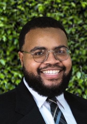 Smiling businessman with beard and glasses wearing a suit and tie in front of a green hedge.