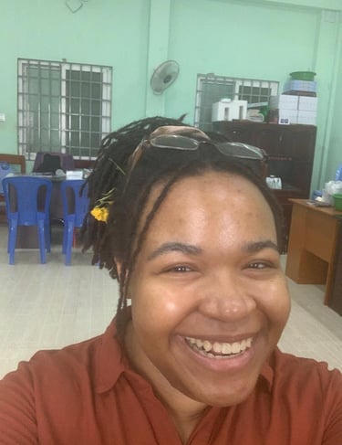 Smiling woman with locs and glasses on her head posing for a selfie in a brightly lit office room.