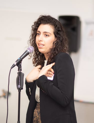 Woman behind microphone, making hand gestures, flipchart in the back, seminar setting.