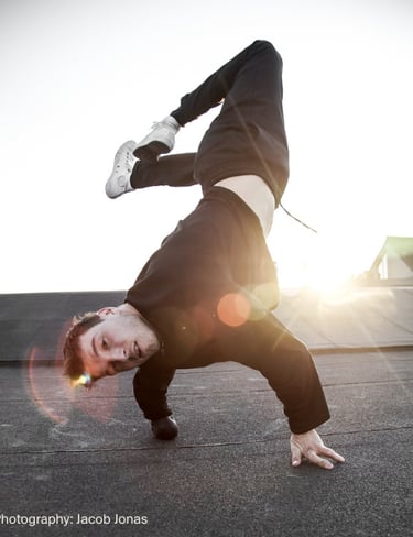 Athletic breakdancer performing a one-handed freeze on a rooftop at sunset with lens flare.