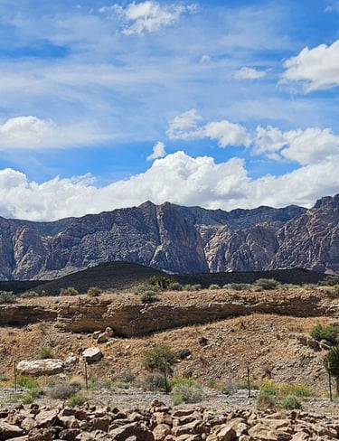 Panoramic view of rugged desert mountains and rock formations under a bright blue sky with white clouds.