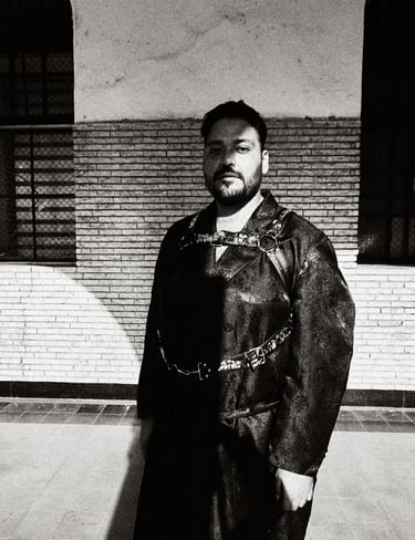 Black and white portrait of a bearded man wearing a leather harness over a coat against a brick wall.