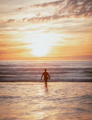 A silhouette of a surfer standing in the ocean, walking into the sun. Vibrant orange and yellow
