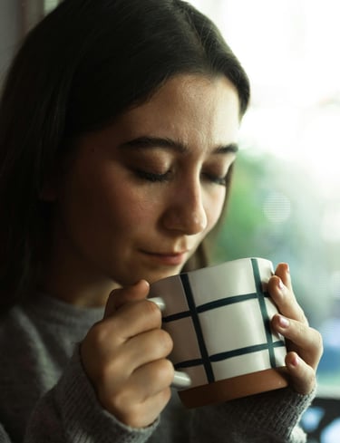 A young woman peacefully holding a ceramic mug while enjoying a hot cup of coffee or tea indoors.