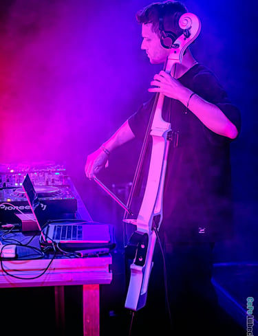 A male musician performs a live electronic set playing an electric cello with DJ equipment under purple stage lights.