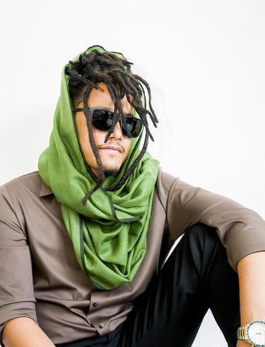 Man with dreadlocks wearing sunglasses and green pashmina scarf, styled with brown shirt and watch.