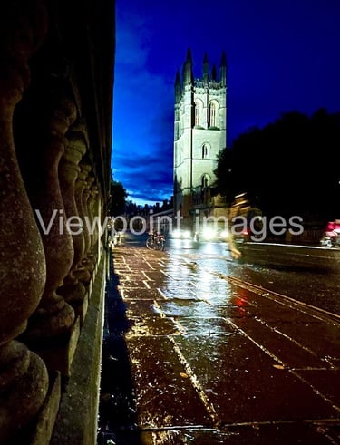 Magdalen College Tower at night viewed from a rainy Oxford street with reflections on wet pavement.