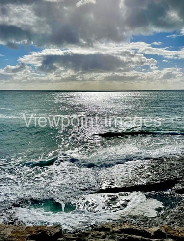 Sparkling blue ocean waves crashing against a rocky shore under a cloudy sky with bright sunlight.